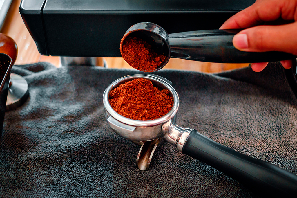 Foto aproximada de uma mão segurando uma colher e derramando pó de café em um portafilter.