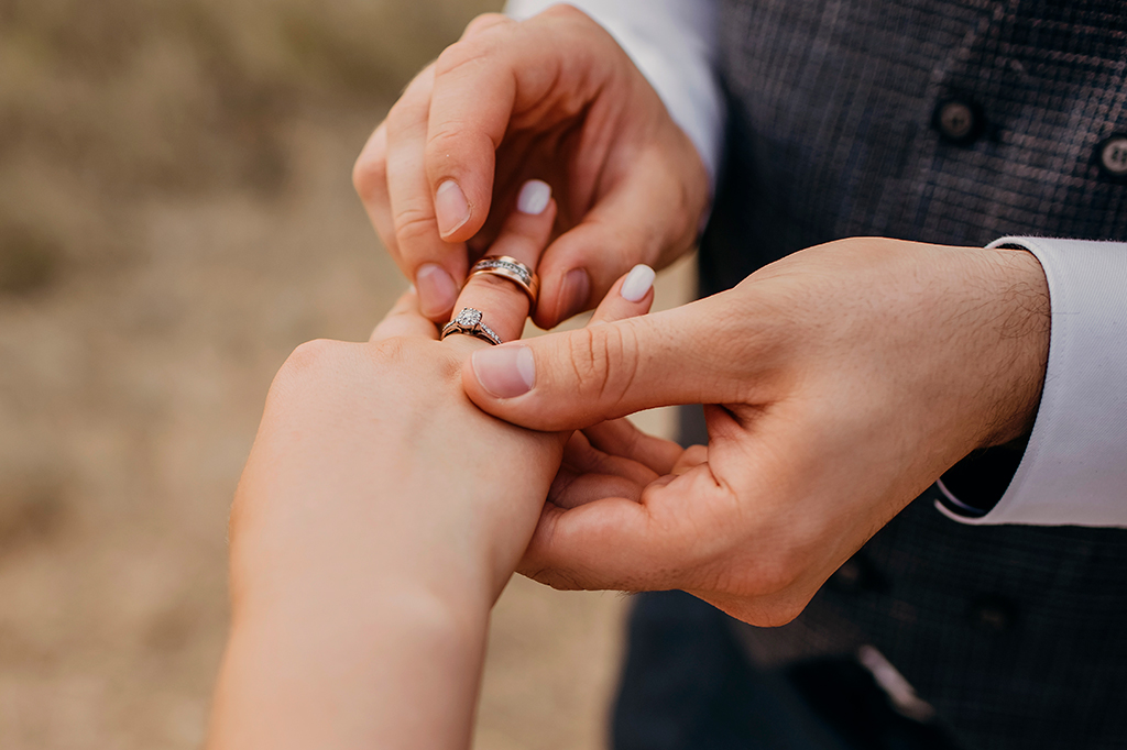 Foto de cerimônia de casamento, foco na mão do noivo colocando a aliança de casamento no dedo da noiva.