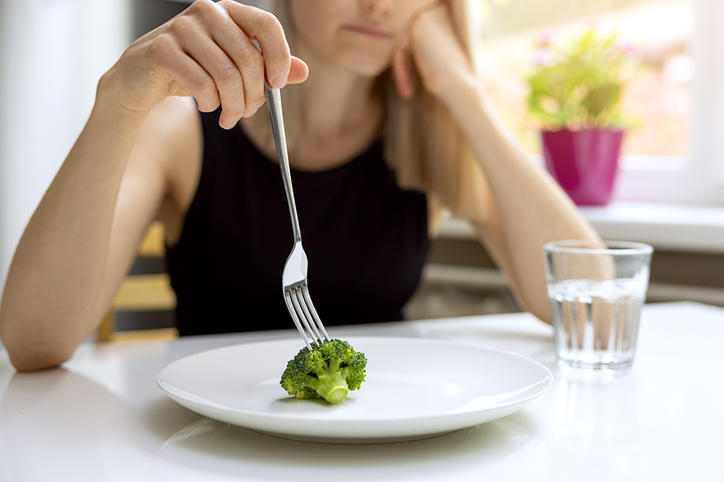Foto aproximada de uma mulher olhando infeliz para o garfo apoiado em cima de um brócolis.