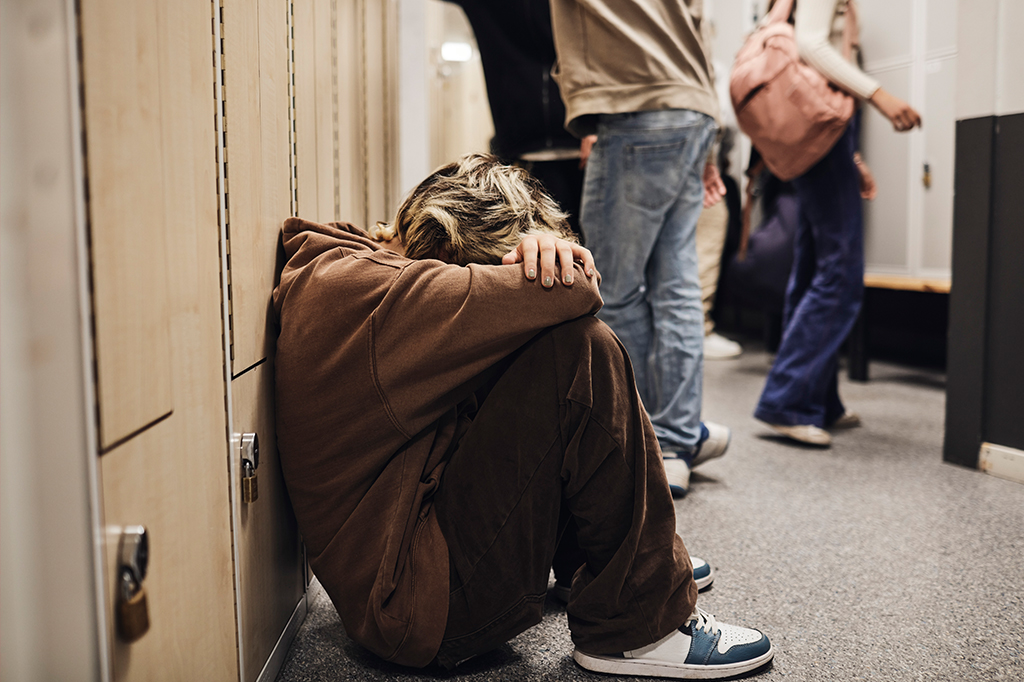 Imagem de um jovem sentado encostado nos armários de um corredor de escola.