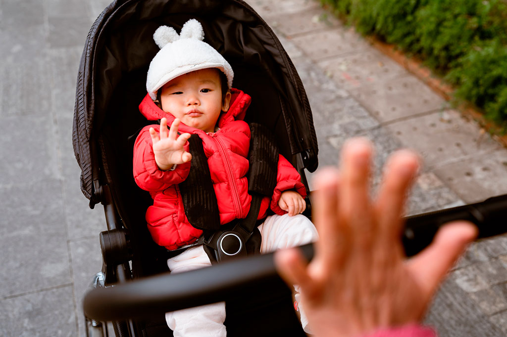 Foto de um bebê dentro do carrinho de passeio, acenando. No canto inferior direito, há uma mão adulta desfocada.