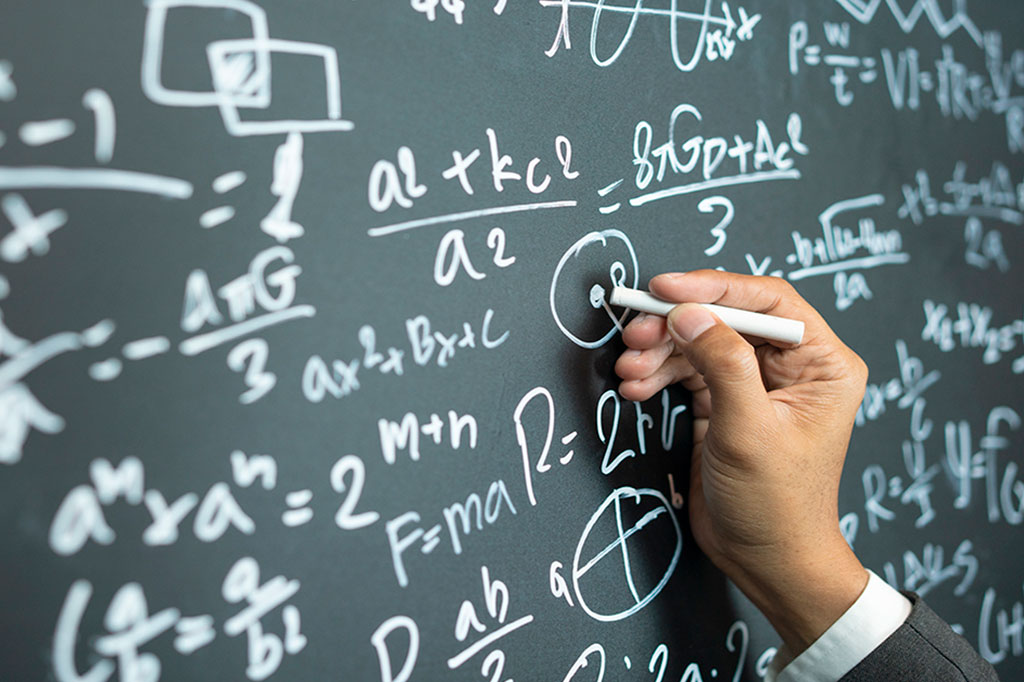 Foto aproximada da mão de um homem escrevendo no quadro negro; no quadro há varias formulas matemáticas.
