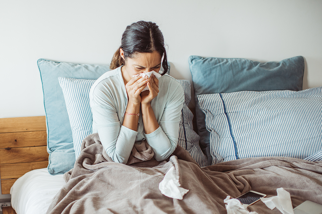 Imagem de uma mulher doente sentada na cama enquanto assoa o nariz.