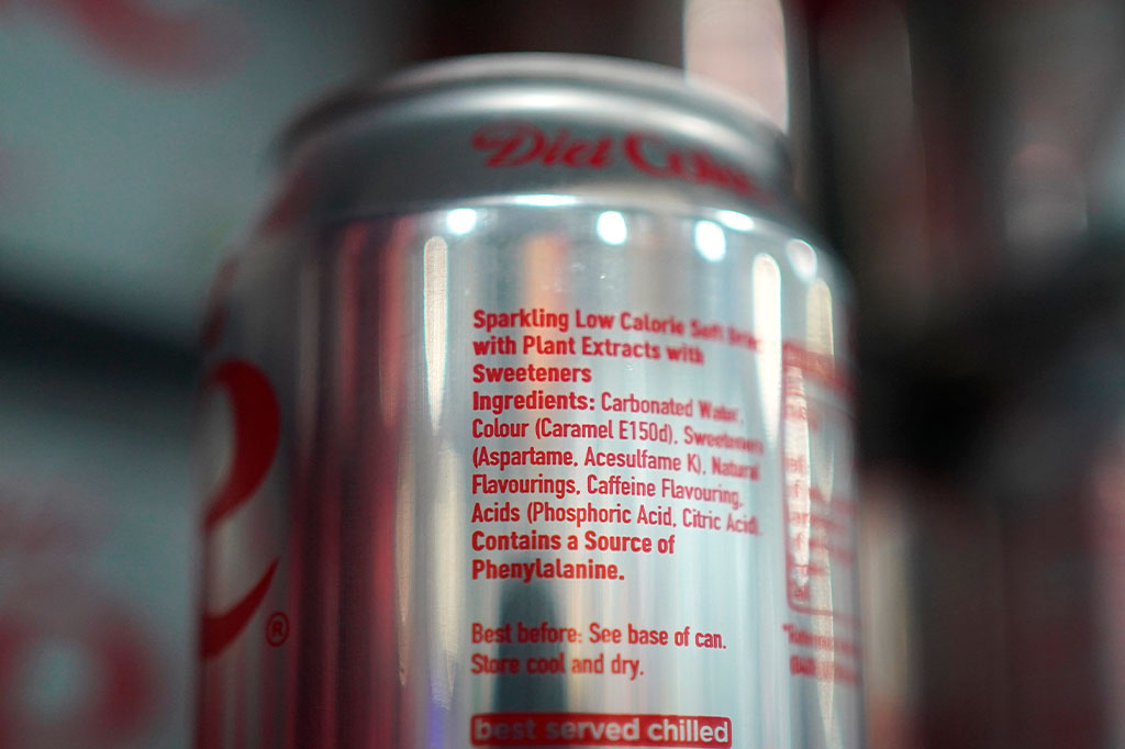 Foto aproximada do rótulo de uma lata de Coca-Cola na prateleira do mercado. Na lista de ingredientes do rótulo podemos ver o uso de Aspartame, que será listado como "possivelmente cancerígeno para humanos" a partir do próximo mês, com base nas descobertas da Agência Internacional de Pesquisa sobre o Câncer (IARC) da Organização Mundial da Saúde (OMS).