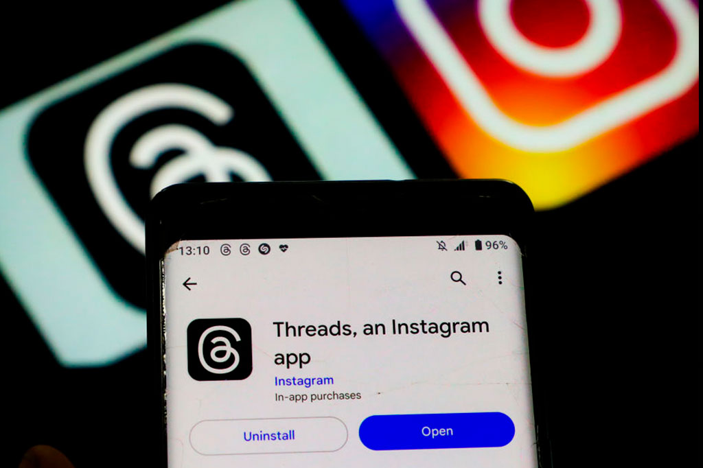 Foto aproximada da tela do celular mostrando o novo aplicativo "Threads" instalado. Ao fundo, o logo do mesmo aplicativo junto ao logo do aplicativo Instagram.
