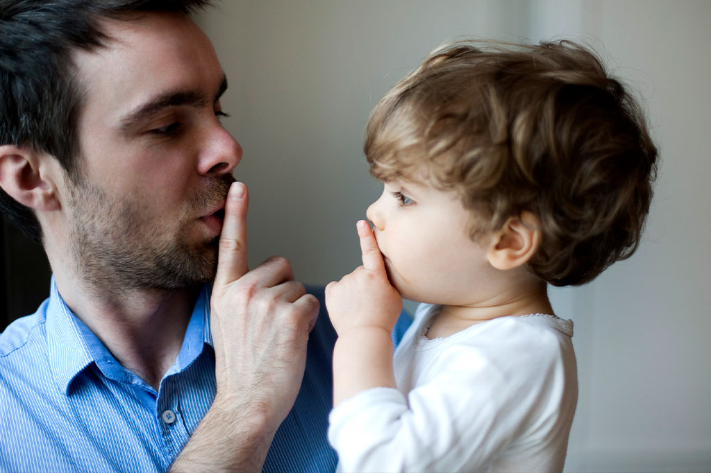 Pai segurando seu filho no colo e o indicando, com o dedo entre os lábios para ficar calado; a criança está repetindo o sinal.