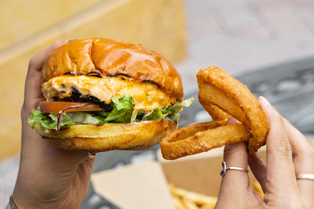 Foto aproximada de uma mão segurando hambúrguer e onion rings.