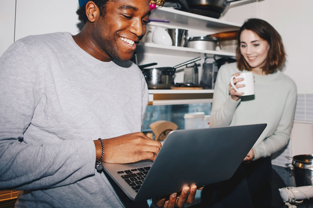 Foto de homem feliz mostrando laptop para amiga, que segura uma xícara de café. Ambos estão na cozinha.