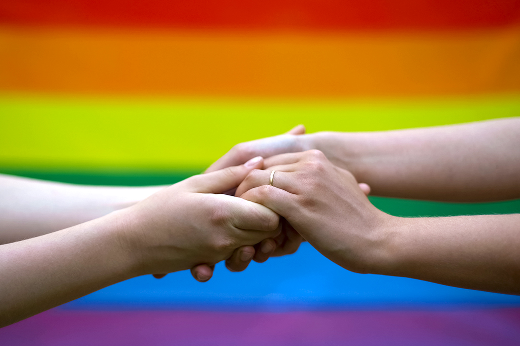 Imagens aproximada de um casal segurando mãos na frente da bandeira LGBTQIAP+.