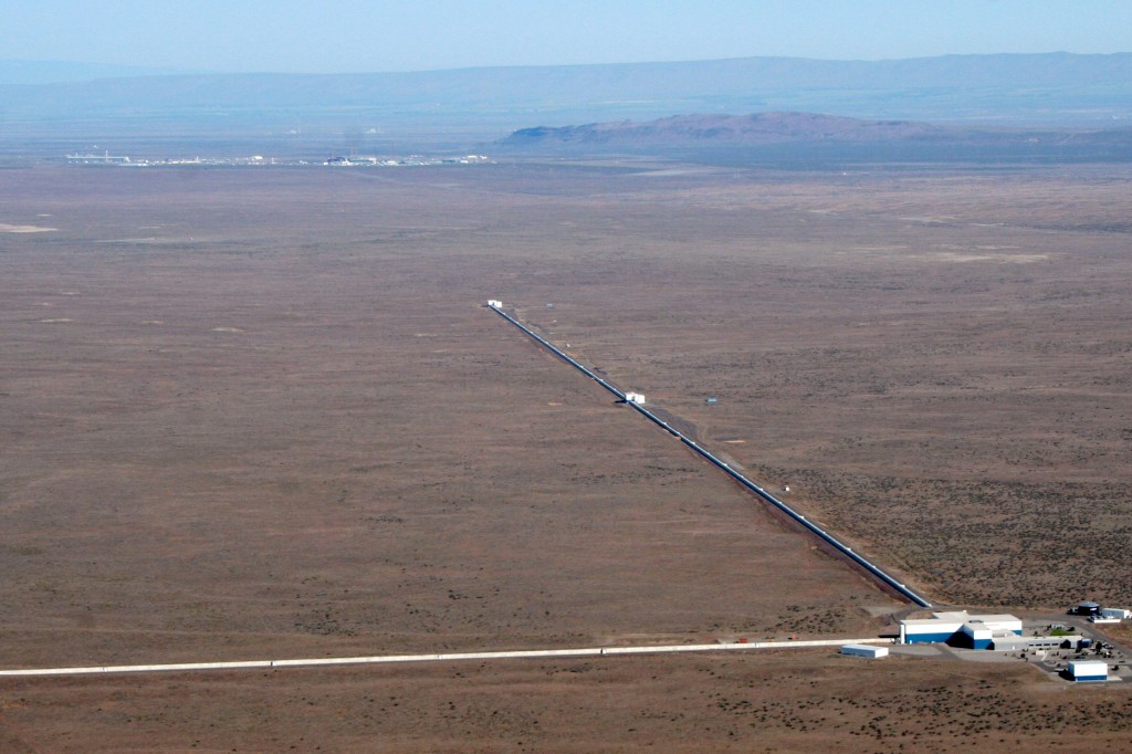 Visão aérea de um dos prédios que compõe o observatório LIGO. Trata-se de um prédio com dois longos braços em forma de L localizado no meio do deserto.