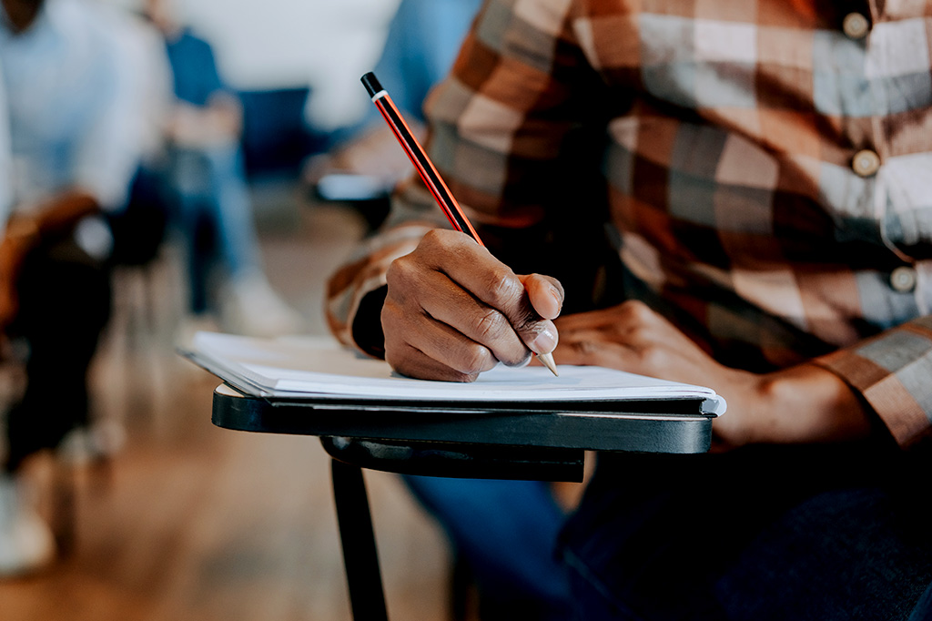 Foto aproximada de uma pessoa em uma sala de aula escrevendo em um papel durante uma prova.