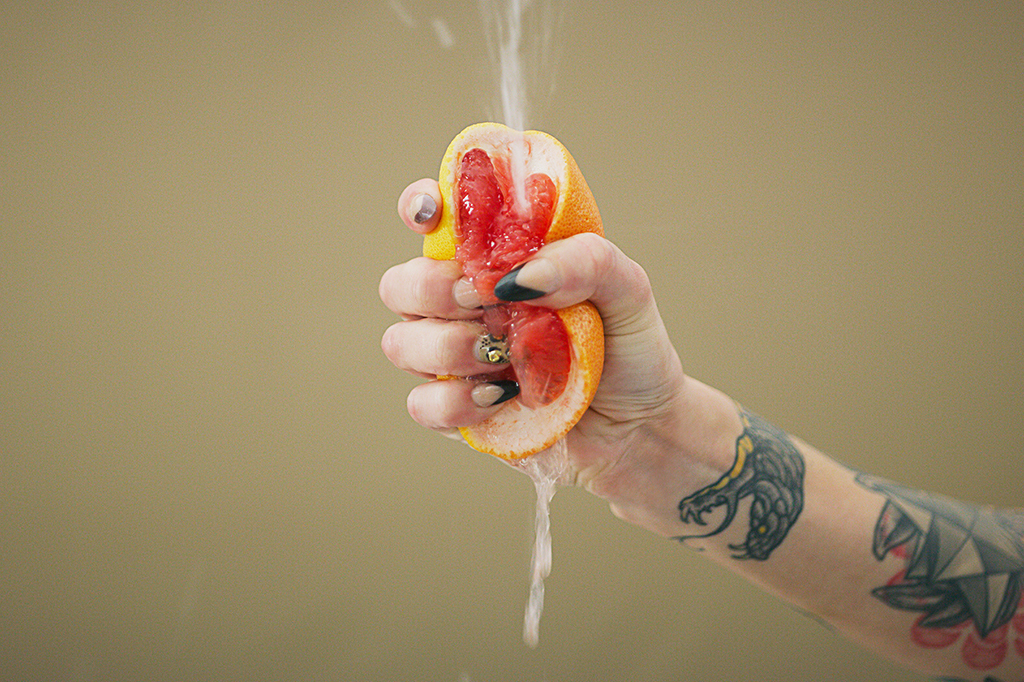 Imagem de uma laranja sendo espremida por uma mão feminina.