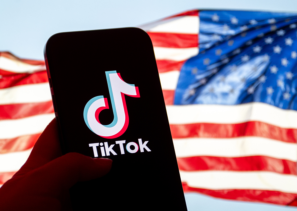 Mão segurando um celular que apresenta o logo do TikTok em sua tela. A bandeira dos Estados Unidos aparece desfocada ao fundo.