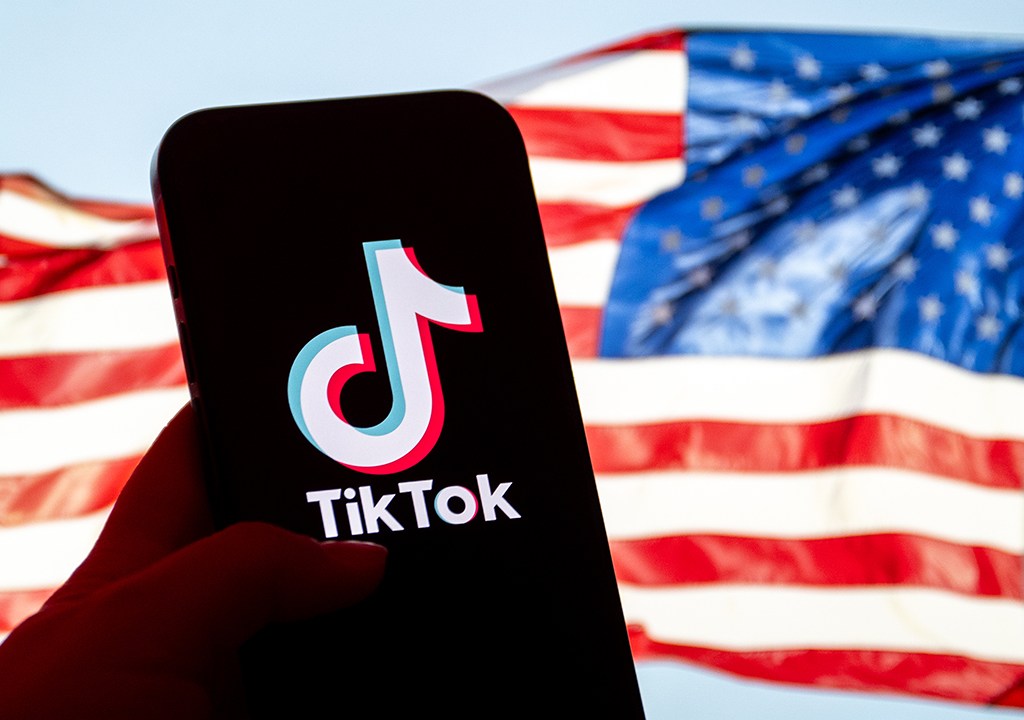 Mão segurando um celular que apresenta o logo do TikTok em sua tela. A bandeira dos Estados Unidos aparece desfocada ao fundo.