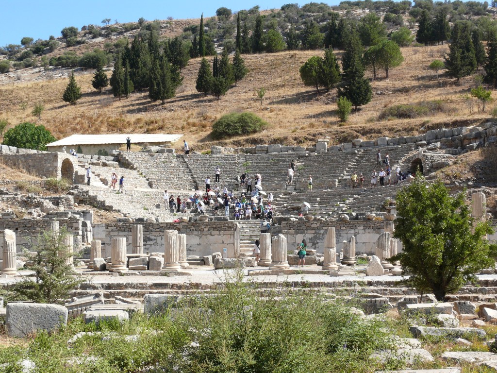 A imagem mostra um antigo anfiteatro de pedra, cinza, sem teto, em ruínas, localizado no sopé de um morro arborizado.
