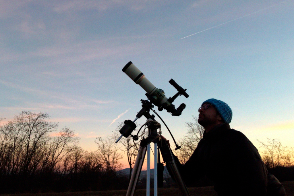 Silhueta de um homem, telescópio e campo sob o céu ao entardecer.