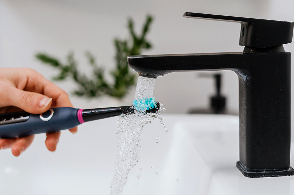 Foto aproximada de uma mão segurando uma escova de dente eletrônica embaixo de uma torneira aberta.