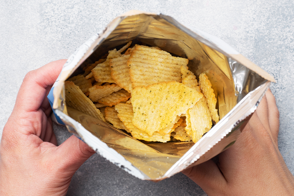 Vista de cima de duas mãos segurando um pacote de batatas chips, aberto, e vê-se o interior prateado.