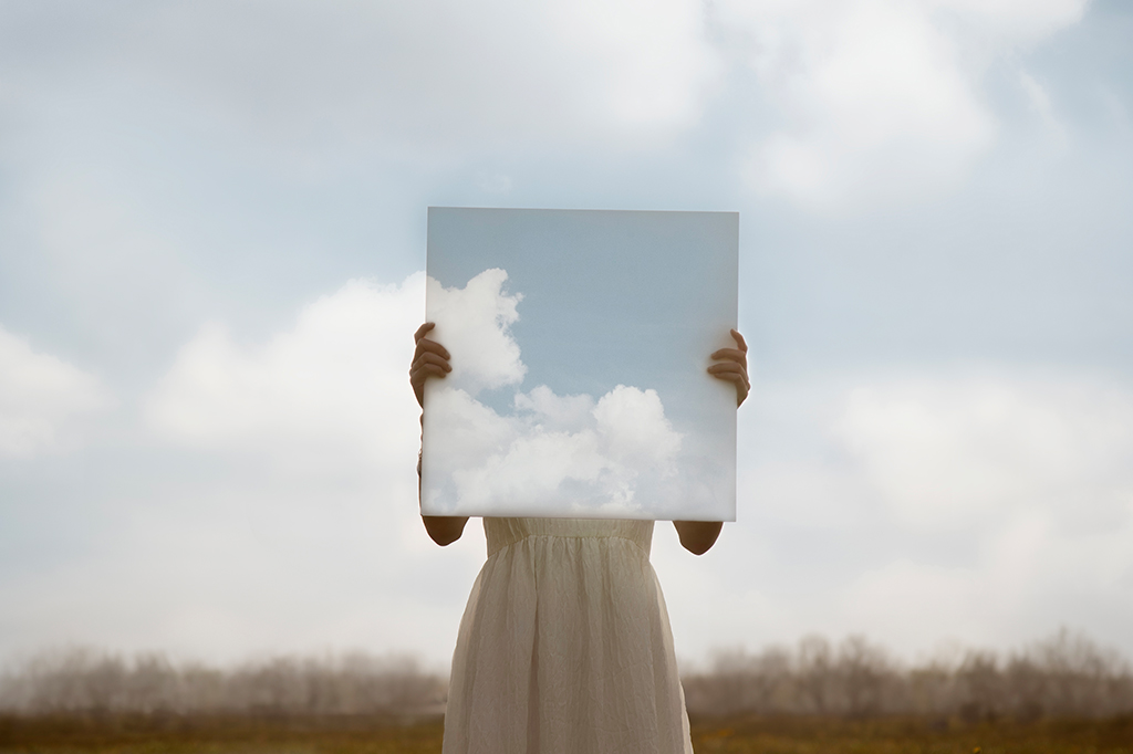 Close up de uma mulher segurando uma pintura surreal do céu na frente de seu rosto, mesclando com o céu em segundo plano.
