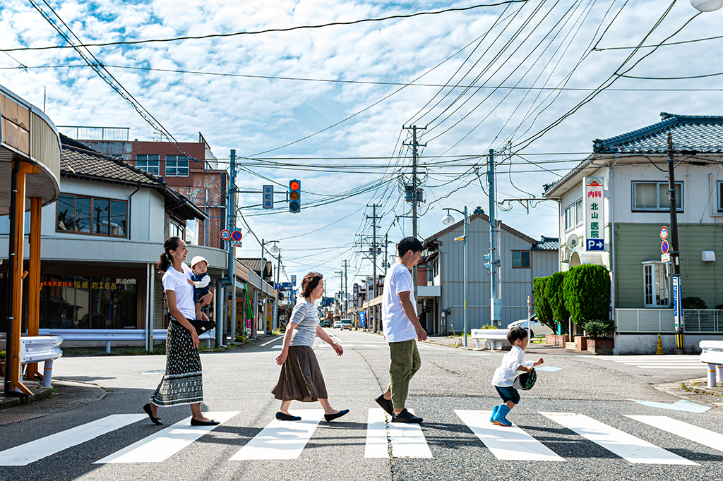 Quatro membros de uma família atravessando uma faixa de pedestres. O cenário é de alguma cidade Japonesa.