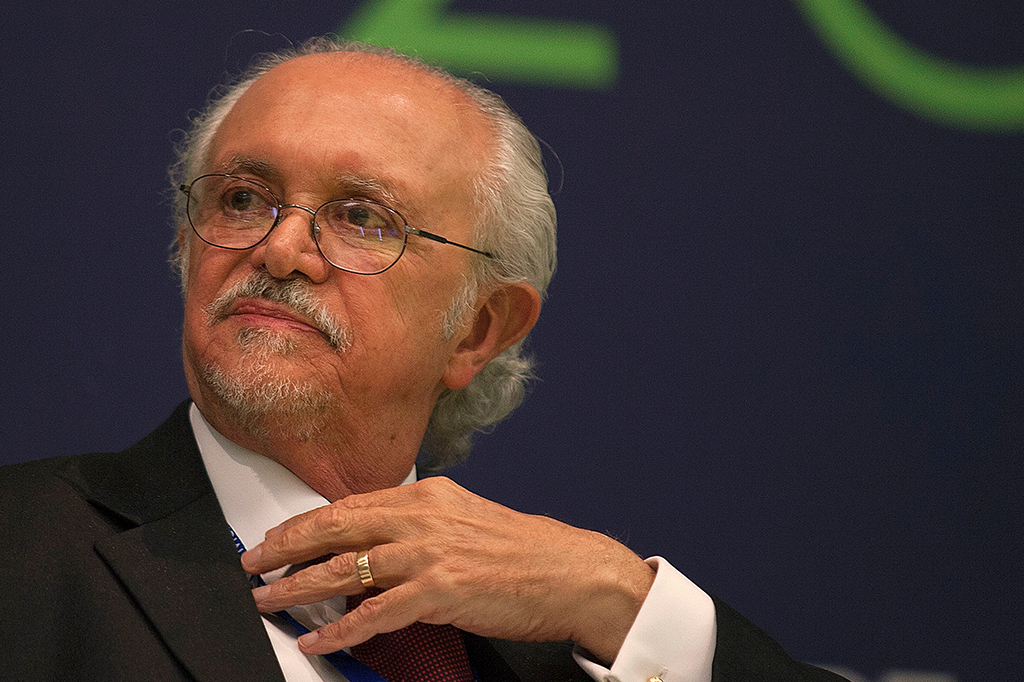 Homem idoso de cabelos brancos, bigode, barba e óculos. Ele veste um terno.