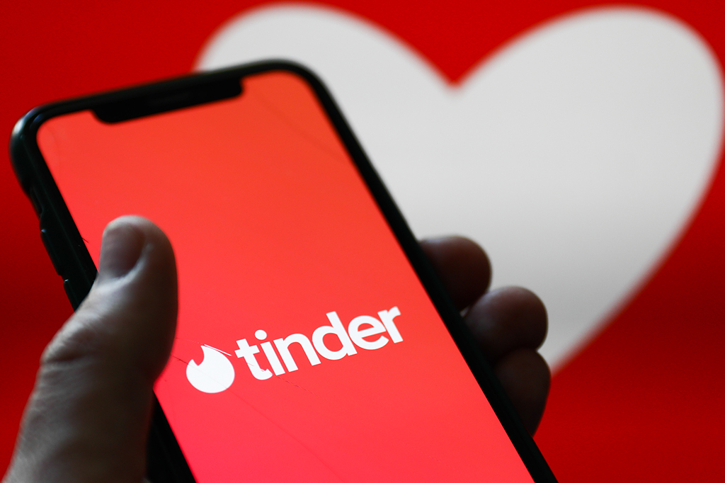 Mão segurando um celular exibindo o logo do aplicativo Tinder. Há um desenho de um coração no fundo.
