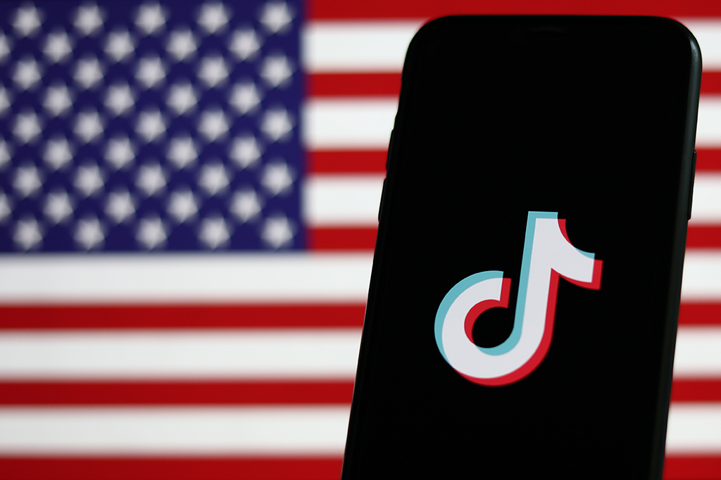 Logotipo do aplicativo TikTok em um celular. Ao fundo, há uma bandeira dos Estados Unidos.