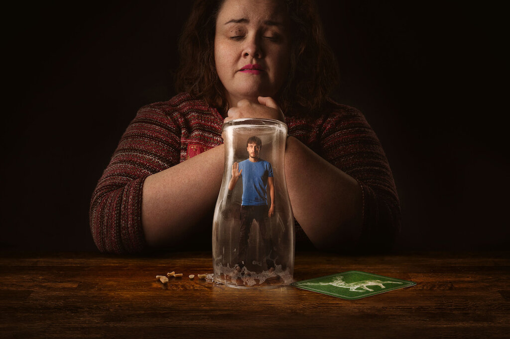 Uma mulher com os braços apoiados em uma mesa de madeira agarrando suas próprias mãos de olhos fechados. Em cima da mesa há um copo de vidro virado para baixo com a miniatura de um homem dentro do recipiente.