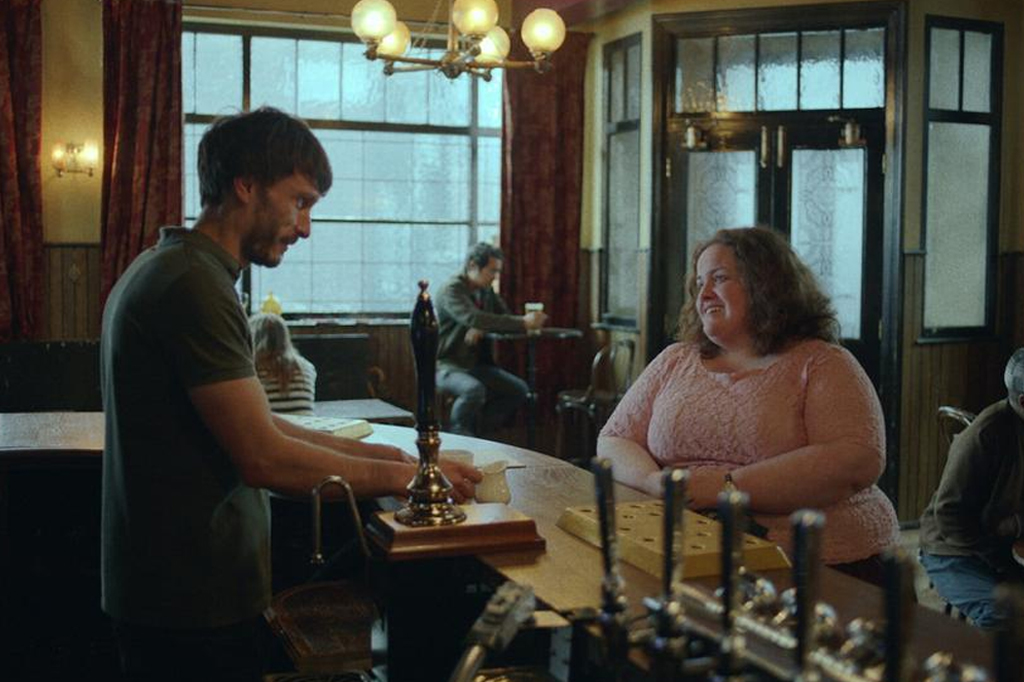 Um homem em pé na parte interna de um balcão de um bar. Há uma mulher sentada, do lado externo, em uma banqueta.