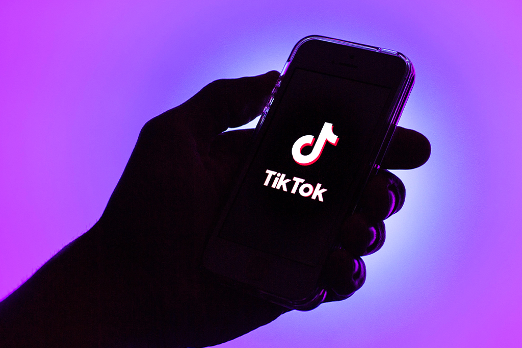 Foto contra luz de uma mão segurando um celular com logo do Tik Tok à mostra.