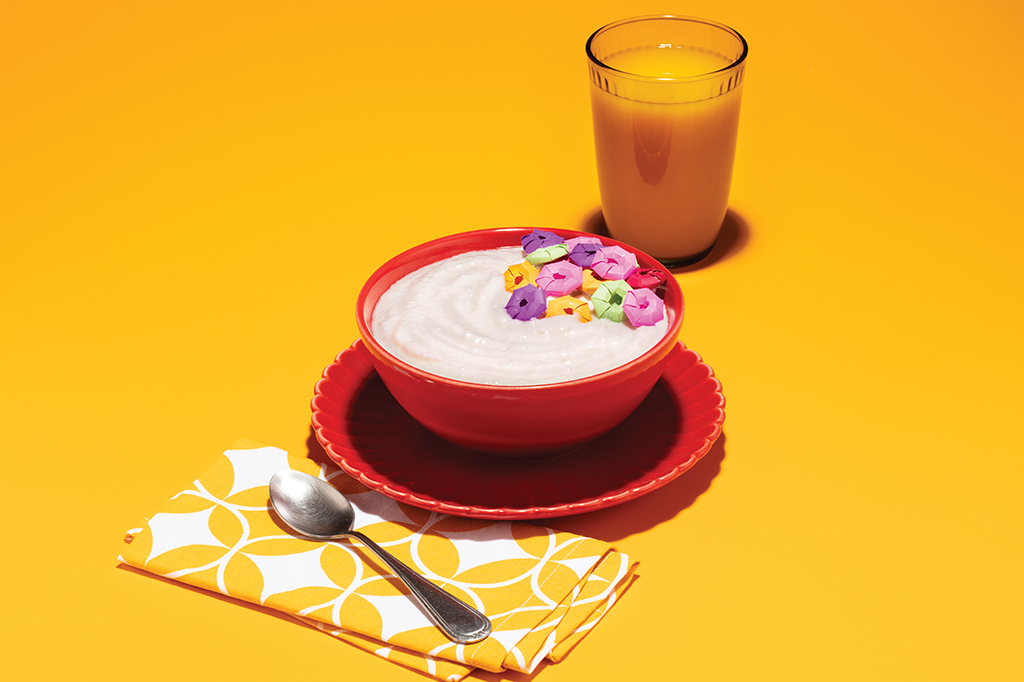 Foto de uma tigela com leite e cereais coloridos feitos de papéis ocupando 1/4 dela, envolta dela tem um guardanapo de tecido com colher em cima e um copo com suco.