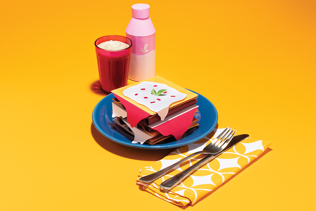 Foto de um prato com uma lasanha feita de papel, do lado um guardanapo de tecido e talheres em cima e um copo com iogurte com uma embalagem de iogurte feita de papel.