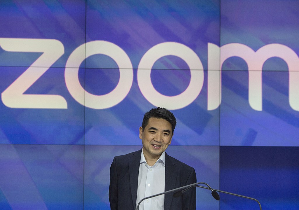 Homem asiático vestindo roupa social. No fundo, há o logo da empresa "Zoom".