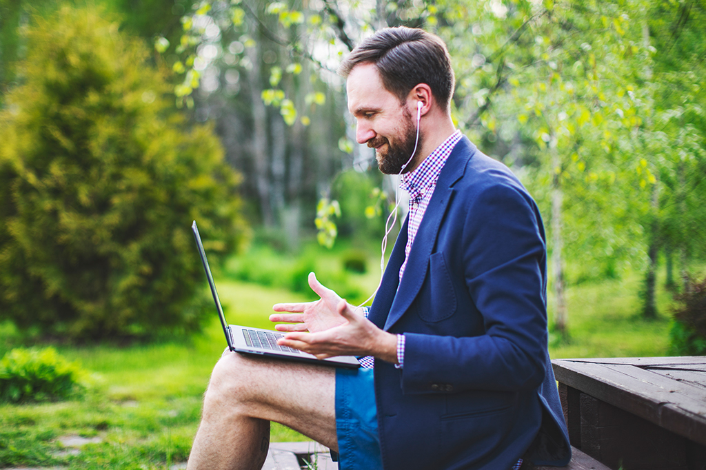 Um homem de terno sentado em um degrau de madeira vestindo terno e bermudas. Ele está com um notebook apoiado em seu colo ouvindo fones de ouvido.