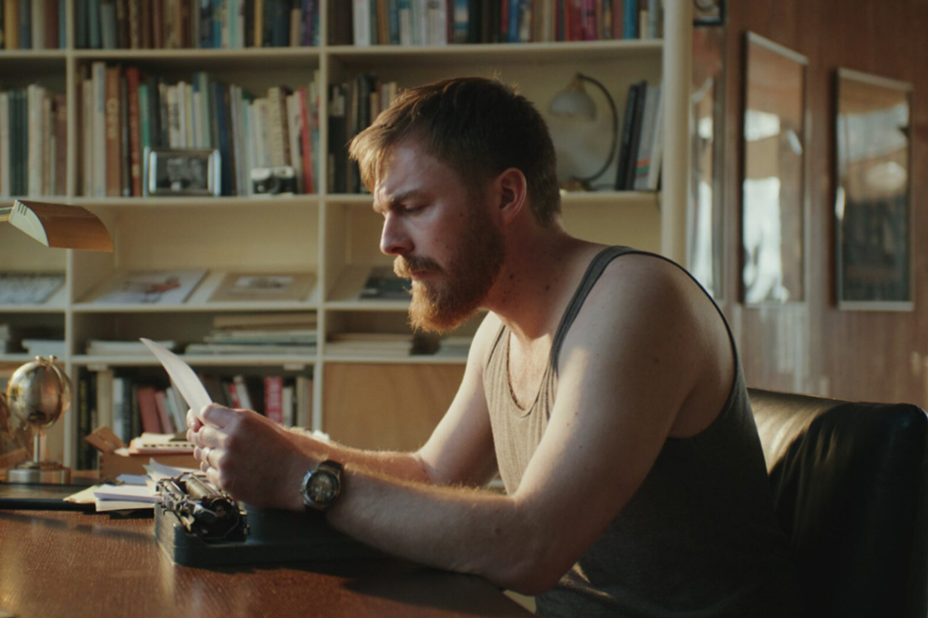 Homem de blusa regata sentado diante uma mesa segurando um papel em suas mãos.
