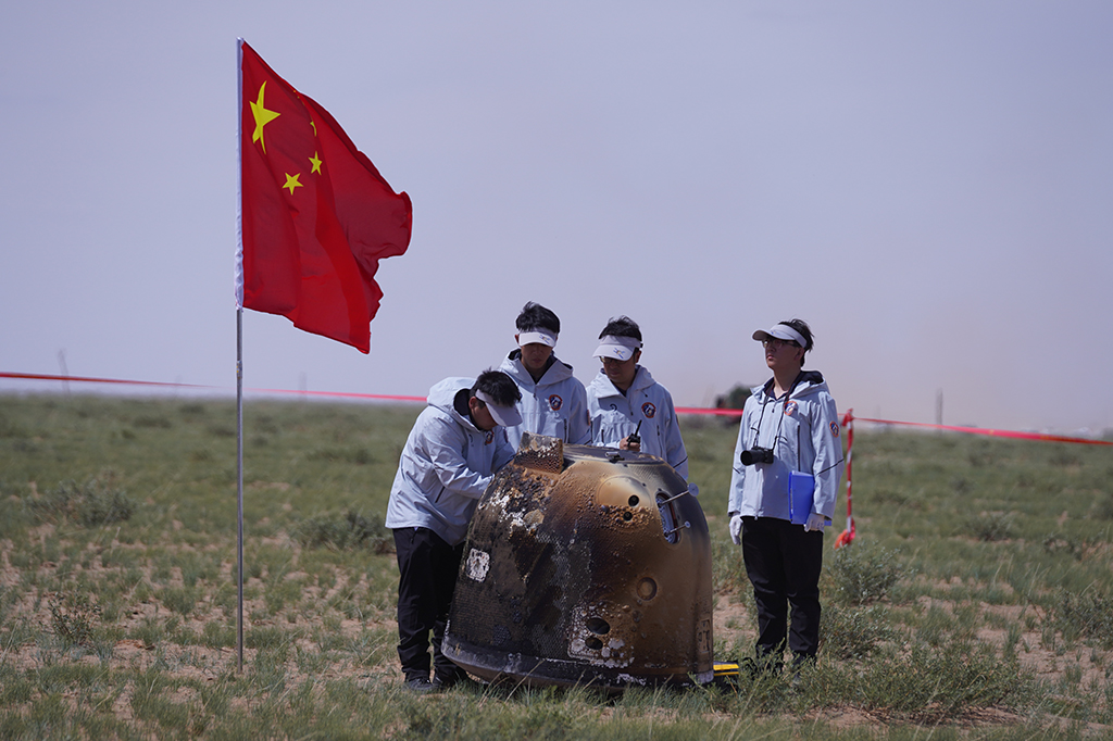 A cápsula de retorno da sonda Chang'e-6 pousando na área de pouso. Há uma bandeira da China ao lado e quatro pessoas no entorno.