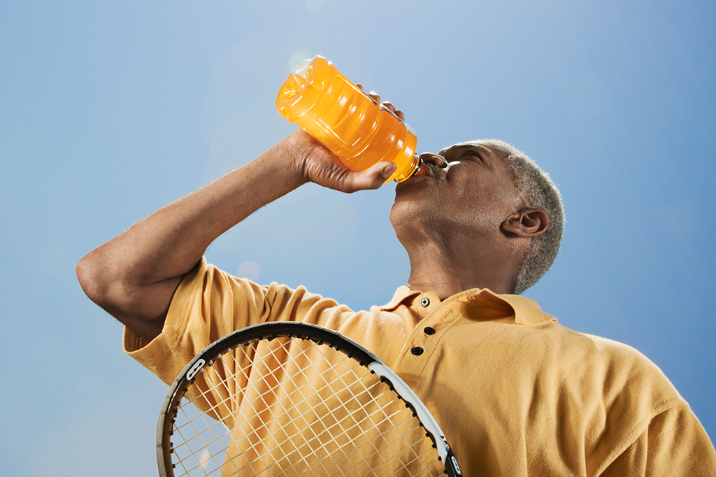 Um homem bebendo uma bebida de cor amarela em uma mão e, na outra, segurando uma raquete de tênis.