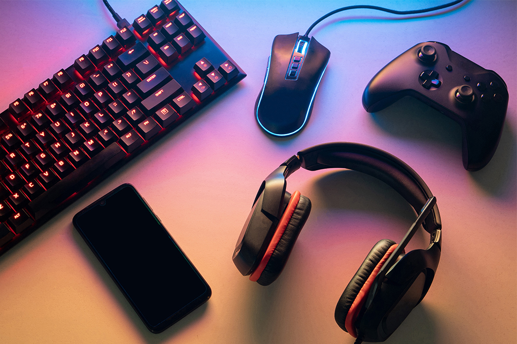 Imagem de diversos equipamento de jogos em cima de uma mesa, sendo eles um teclado, mouse, controle, fone de ouvido e celular.