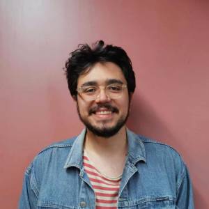 foto Foto de um homem branco com barba preta, cabelos pretos, camiseta listrada vermelha e branca, jaqueta jeans azul clara em um fundo rosa.