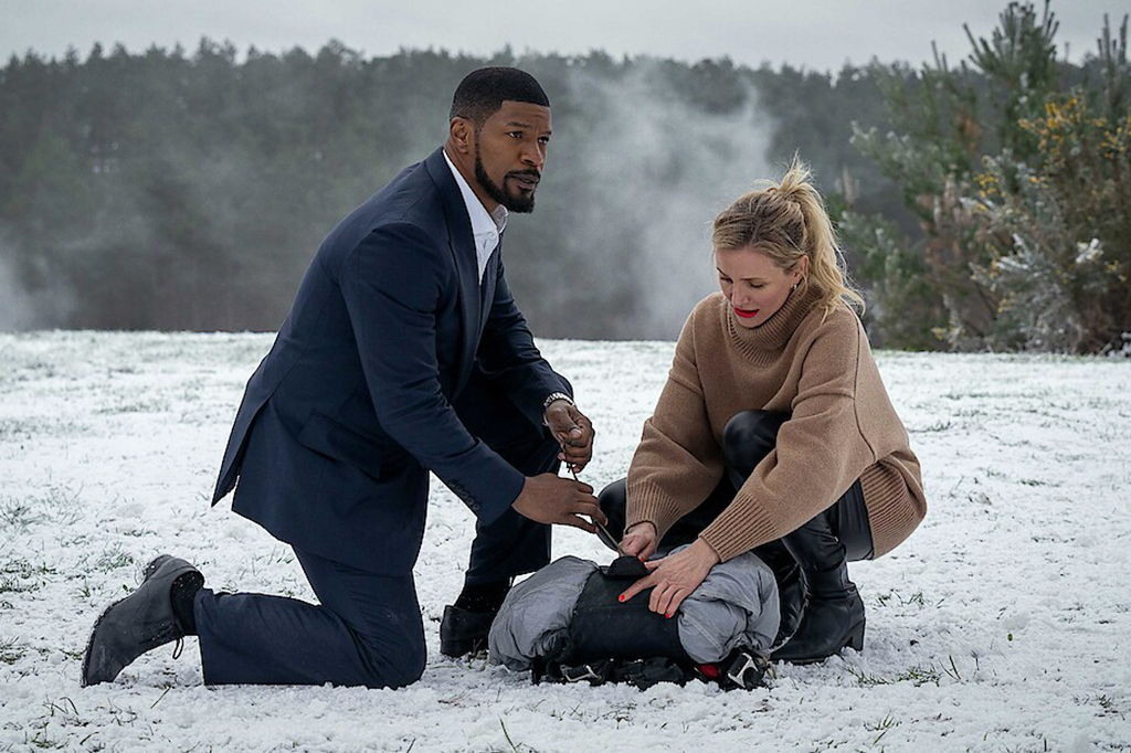 Um homem ajoelhado, vestido de terno. Ao seu lado direito há uma mulher, de roupas de frio. Ambos estão mexendo em uma mala. O cenário é de uma vegetação e neve.