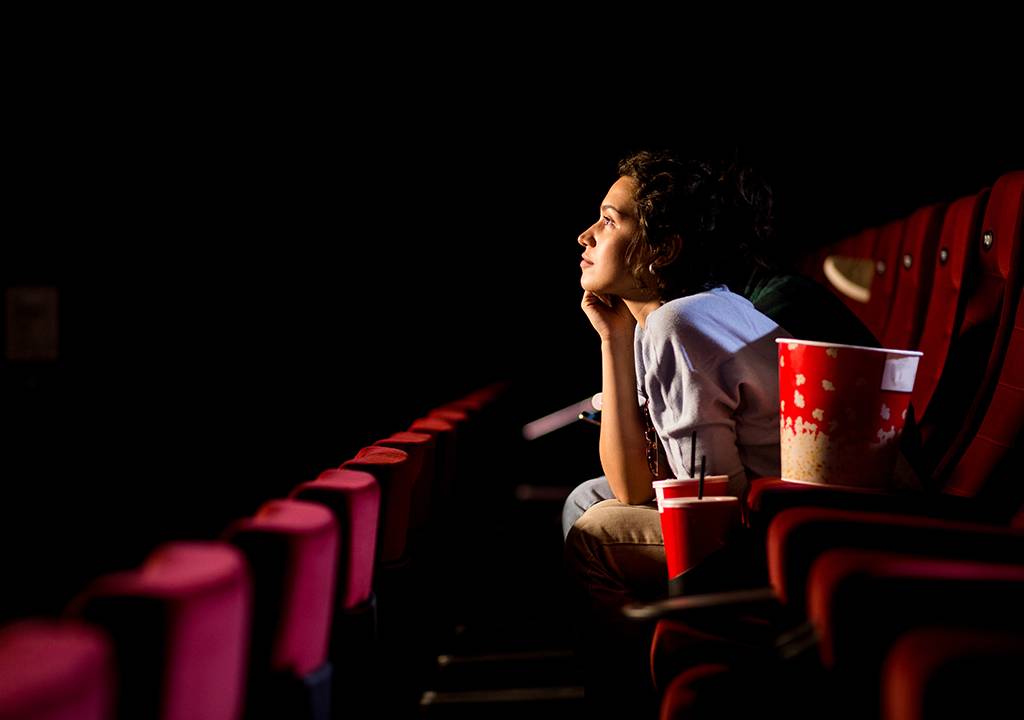 Uma mulher assistindo a um filme sozinha no cinema. Ao seu lado, há copos e um grande balde de pipoca.