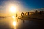 Pôr-do-sol em uma praia. Há pessoas jogando futebol na areia.