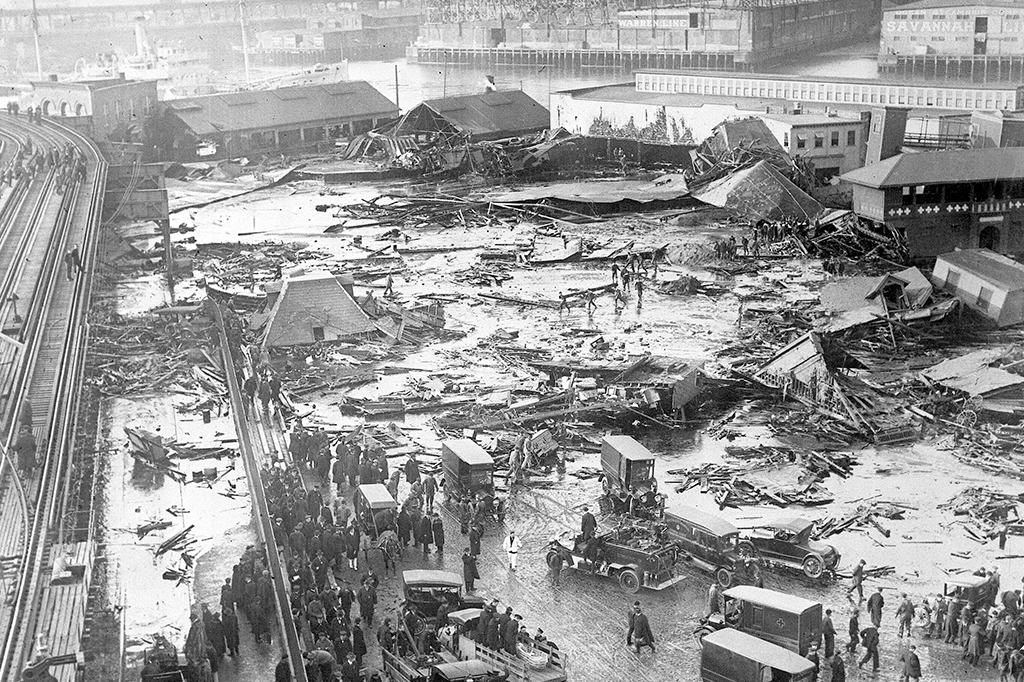 Fotografia Destroços do tanque desabado visíveis ao fundo, ao centro, próximo ao armazém de cor clara. Estrutura ferroviária elevada visível na extrema esquerda e a praia balnear do North End Park na extrema direita.