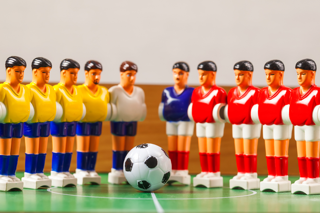 Imagem de bonecos de jogo de pebolim perfilados diante de uma bola de futebol.