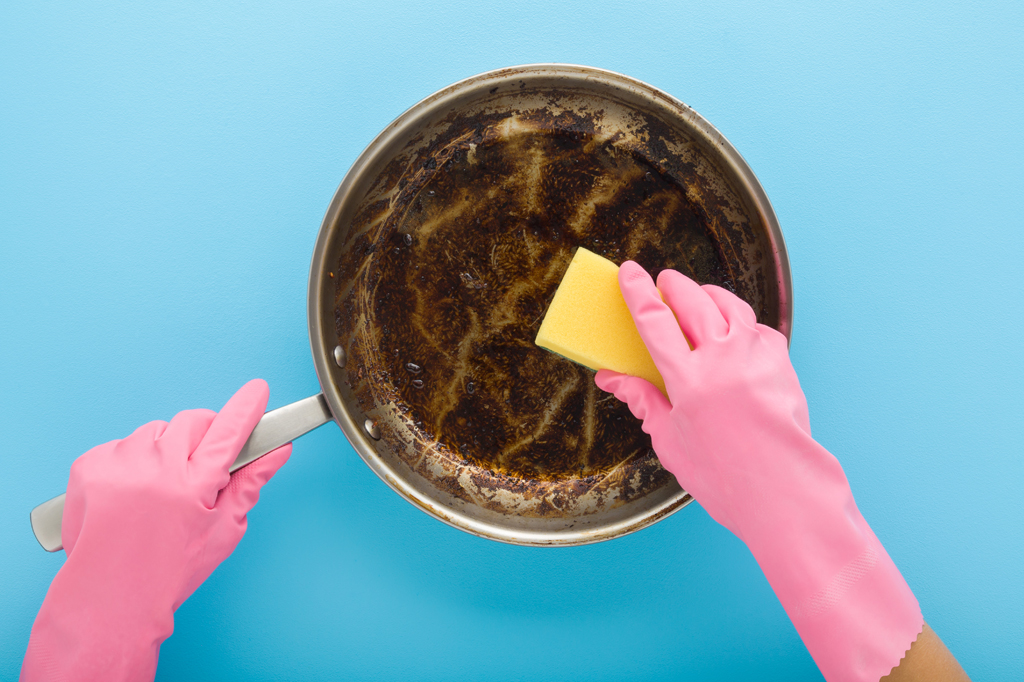 Imagem, em fundo azul, de duas mãos protegidas por luvas de borracha rosa, segurando uma esponja amarela e lavando uma frigideira de aço inoxidável com mancha de gordura queimada.