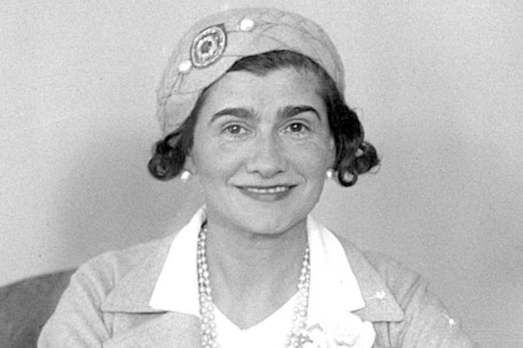Image de uma mulher sorrindo, usando chapéu, colar e brincos de pérolas.