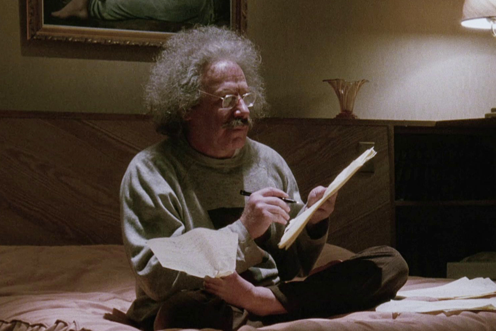 Imagem de um homem idoso sentado na cama escrevendo num caderno.