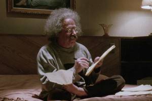 Imagem de um homem idoso sentado na cama escrevendo num caderno.