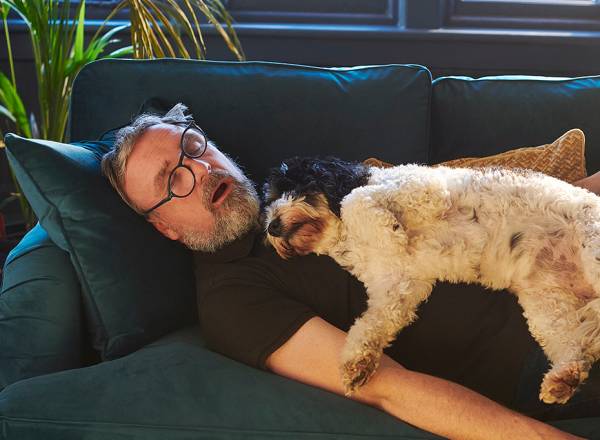 Fotografia de um homenm dormindo no sofá com o seu cachorro.