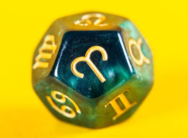 Imagem, em fundo amarelo, de dado de astrologia verde com símbolo do zodíaco de Áries em destaque.